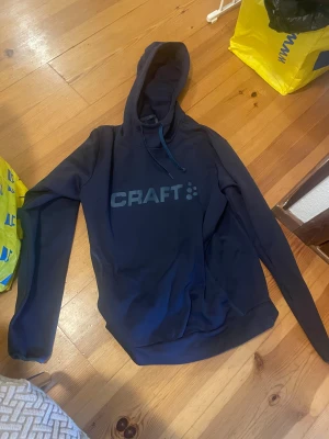 Mörkblå hoodie - Snygg mörkblå hoodie från Craft i storlek S. Tröjan har huva med snörning och stort Craft-tryck på bröstet. Tillverkad i mjukt syntetmaterial, perfekt för träning eller chill. Långärmad och med klassisk passform.