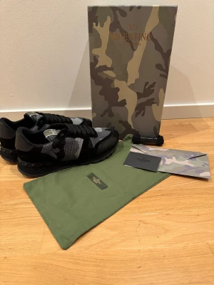 Valentino Garavani Rockrunner sneakers - Snygga Valentino Garavani Rockrunner sneakers i svart och grått med camouflage-mönster. Skorna har snörning, platt sula med nitar och detaljer i mocka och mesh. Märket syns tydligt på plösen och innersulan. Perfekt för dig som gillar exklusiv streetstyle. Passar även storlek 43. Priset kan diskuteras vid snabb affär!