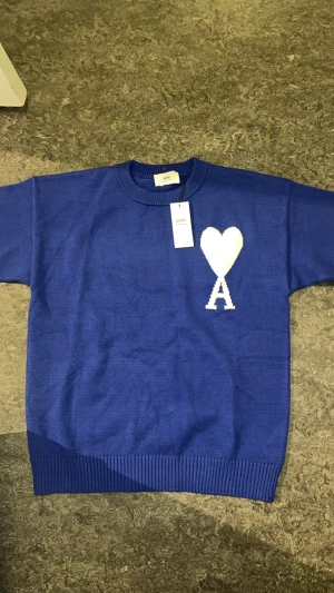 Blå stickad tröja från Ami - Blå stickad tröja från Ami Alexandre Mattiussi med rund hals och ribbade muddar. På bröstet finns ett vitt hjärta med bokstaven A, vilket ger en unik och snygg detalj. Perfekt för dig som gillar stilrena och coola plagg.                                              Inte använd.