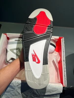 Nike Air Jordan 4 White Cement - Nike Air Jordan 4 White Cement med vit ovandel i skinn, grå och svarta detaljer samt specklad grå sula. Klassisk meshpanel på sidorna, svart Nike Air-logga på hälen och röd Jumpman på plösen. Snygg och ikonisk siluett som sticker ut.