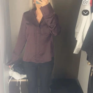 Mörklila satinblus med knappar - Säljer en stilren mörklila blus i satin med klassisk krage och knappar framtill. Skjortan har lång ärm och en lätt glansig finish som ger en lyxig känsla. Perfekt för dig som gillar en clean och elegant look.