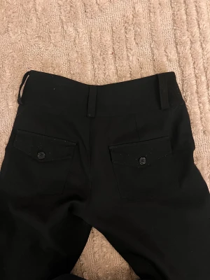 Svarta bootcut kostym byxor med fickor från Gina Tricot  - Svarta bootcut kostym byxor med två knappar fram och praktiska fickor med lock och knapp bak. Byxorna har bälteshällor och är sydda i ett slätt tyg som ger en clean look. Passar dig som gillar klassisk stil med lite edge.