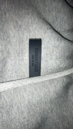 Grå Essentials mjukisbyxor med snörning - Grå mjukisbyxor från Essentials Fear of God med elastisk midja och långa snören framtill. Byxorna har ribbade muddar vid bensluten och logotyptryck på vänster ben. Perfekt för chill och streetwear. Materialet är mjukt och bekvämt bomull.