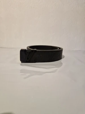 Svart Louis Vuitton skärp i skinn - Snyggt svart bälte från Louis Vuitton med klassiskt LV-spänne och diskret mönster längs hela remmen. Tillverkat i skinn och passar perfekt för att lyfta vilken outfit som helst med en exklusiv touch.