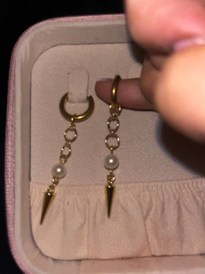 Guldiga örhängen med pärlor och spikes - Säljer ett par trendiga örhängen i guldfärgad metall med hängande detaljer. Örhängena har små ringar, vita pärlor och spetsiga spikes längst ner. Perfekta för dig som gillar en edgy och modern stil.