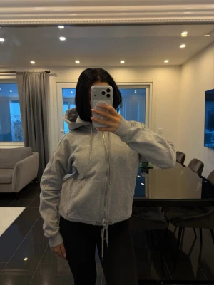 Grå hoodie med dragkedja från Nicki studios - Grå zip-hoodie från Ronnie Mind med huva och dragsko i midjan. Hoodien har fickor framtill och ribbade muddar vid ärmslut. Perfekt för en chill och avslappnad stil.