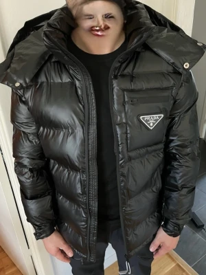 Svart dunjacka från Prada med huva - Snygg svart dunjacka från Prada med glansig finish, stor huva och dragkedja framtill. Jackan har en bröstficka med dragkedja och ikonisk Prada-logga på vänster sida. Perfekt för kalla vinterdagar och riktigt trendig look. Lite mindre i storleken