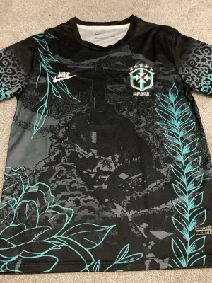 Nike Brasil svart träningströja - Svart träningströja från Nike med Brasil-logga på bröstet. Tröjan har unika turkosa blommönster och detaljer, samt ett stort tryck av Kristusstatyn och stadsmotiv. Ärmarna har leopardmönster. Perfekt för dig som vill sticka ut på träningen.