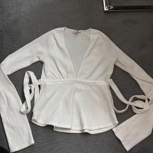 Vit omlottopp med knytband - Säljer en stilren vit omlottopp med långa ärmar och knytband i midjan. Toppen har en v-ringad urringning och är tillverkad i ett lätt, ribbat material. Aldrig använt 
