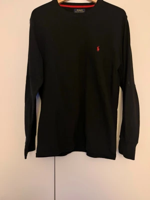 Svart stickad tröja från Polo Ralph Lauren - Klassisk svart stickad tröja från Polo Ralph Lauren med lång ärm och rund halsringning. Tröjan har den ikoniska röda logotypen broderad på bröstet och är tillverkad i ett mjukt, strukturerat material som passar perfekt till alla tillfällen.
