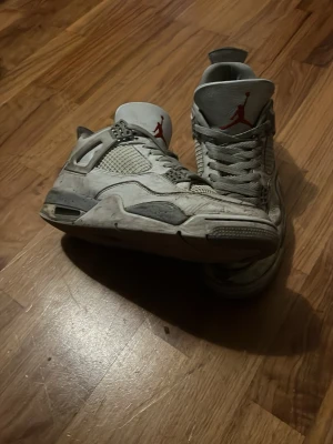 Vita Air Jordan 4 Retro sneakers - Säljer ett par vita Air Jordan 4 Retro sneakers med grå och svarta detaljer samt röd Jumpman-logga på plösen. Skorna har meshpaneler på sidorna och klassisk snörning. Perfekta för dig som gillar ikoniska basketskor med streetwear-vibe.