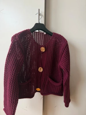 Vinröd virkad kofta med guldknappar - Snygg vinröd virkad kofta med tre stora guldknappar framtill och två fickor. Koftan har en luftig, öppen stickning och rund halsringning. Perfekt för lager på lager och ger en trendig touch till din outfit.