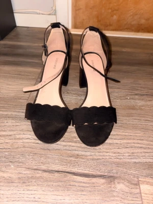 Svarta klackskor  - Svarta klackskor från Graceland. Skorna har en vågig kant framtill och är gjorda i mockaimitation. Klackhöjd på ungefär 8 cm. Använda 1 gång. Utan box.