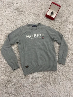 Grå tröja från Morris Rowing Club - Snygg grå tröja från Morris Rowing Club med vit text och klubbmärke på bröstet. Tröjan har rund hals, långa ärmar och ribbade muddar vid ärmslut och nederkant. Perfekt för en stilren och avslappnad look. OBS: Klockan medföljer ej