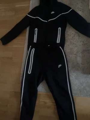 Nike svart träningsset med reflexdetaljer - Säljer ett svart träningsset från Nike med vita reflexdetaljer på både jacka och byxor. Setet har dragkedjor på fickorna och en huva på jackan. Perfekt för dig som vill sticka ut med snygga detaljer när du tränar utomhus.