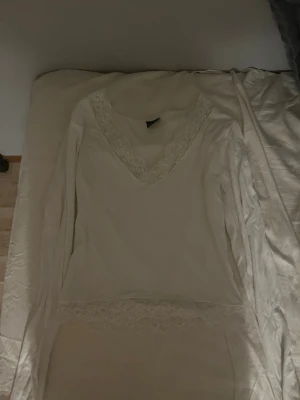 Vit långärmad topp med spetsdetaljer - Söt vit långärmad topp med v-ringning och spetsdetaljer vid halsringningen och nederkanten. Perfekt att styla med jeans eller kjol för en fräsch look. Tunn och mjuk i materialet, passar till många olika outfits.