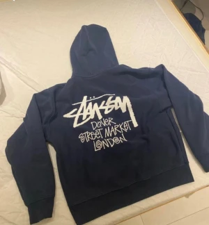 Stüssy x Dover Street Market hoodie - Mörkblå hoodie från Stüssy i samarbete med Dover Street Market London. Stor vit logga på ryggen och mindre tryck på bröstet. Klassisk streetwear-stil med huva och ribbade muddar. Perfekt för dig som gillar exklusiva collabs och snygg design.