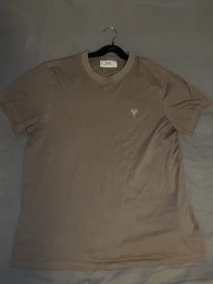 Brun t-shirt från Ami Paris - Säljer en stilren brun t-shirt från Ami Paris med diskret broderad logga på bröstet. T-shirten har rund halsringning och korta ärmar. Tillverkad i mjuk bomull för en skön känsla och avslappnad passform.