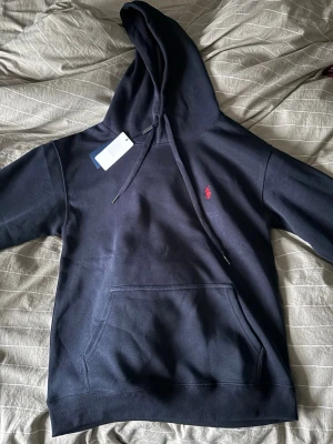 Mörkblå hoodie från Polo Ralph Lauren - Snygg mörkblå hoodie från Polo Ralph Lauren med klassisk röd logga broderad på bröstet. Hoodien har huva med dragsko, magficka och ribbade muddar. Tillverkad i mjuk bomullsblandning som känns skön mot huden. Perfekt för en avslappnad och stilren look.