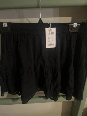 Svart volangkjol med spetskant - Kort svart kjol från Blake med volanger och fin spetskant längst ner. Kjolen har resår i midjan för skön passform och är gjord i ett lätt, vågigt tyg som ger en luftig känsla. Perfekt för dig som gillar en enkel men ändå söt stil.