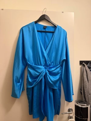 Blå omlottklänning Gina Tricot - Säljer en blå omlottklänning från Gina Tricot med lång ärm och knyt i midjan. Klänningen har v-ringning och en snygg draperad detalj framtill. Perfekt för dig som vill ha en färgstark och trendig look.