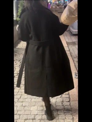 Svart lång kappa från Nelly - ✨Stilren svart kappa från Nelly med lång passform och bälte i midjan. Kappan har klassisk krage och långa ärmar, perfekt för en trendig look. Strl M✨