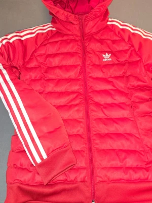 Röd dunjacka med huva från Adidas - Snygg röd dunjacka från Adidas med klassiska vita ränder längs ärmarna och Adidas-logga på bröstet. Jackan har huva, dragkedja framtill och ribbade muddar vid ärmslut och nederkant. Perfekt för kalla vinterdagar och sportig stil.