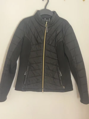 Svart pufferjacka med gulddragkedja - Snygg svart pufferjacka med guldfärgad dragkedja framtill och hög krage. Jackan har quiltade partier och insydda stretchpaneler på sidorna och ryggen för extra komfort. Två fickor med dragkedja och smal passform. Perfekt för kalla vinterdagar.