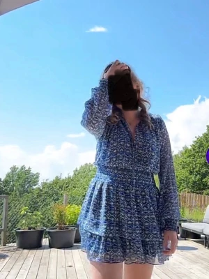 Blå blommig kortklänning med volang - Supersöt blå kortklänning med blommigt mönster och volangdetaljer. Klänningen har långärmade, transparenta ärmar och markerad midja. Perfekt för sommarens alla tillfällen och ger en fräsch och trendig look.