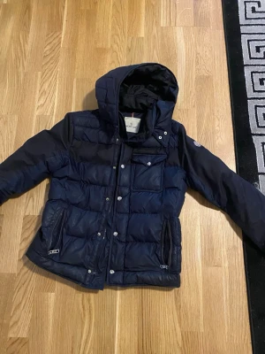 Mörkblå dunjacka från Moncler - Snygg mörkblå dunjacka från Moncler med huva och quiltad design. Jackan har dragkedjor på fickorna, tryckknappar framtill och en bröstficka med lock. Klassisk Moncler-logga på ärmen och varm vaddering för kalla dagar.