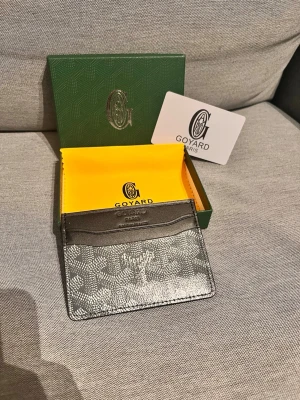 G korthållare i svart och grått - Snygg korthållare från G i svart och grått med klassiskt mönster och präglad logga. Tillverkad i läder och canvas, har flera kortfack och levereras med originalask och putsduk. Perfekt för dig som vill ha stilren och exklusiv accessoar.