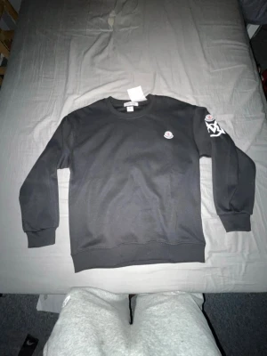 Svart sweatshirt från Moncler - Snygg svart sweatshirt från Moncler med rund halsringning och ribbade muddar. Tröjan har Moncler-logga på bröstet och ett vitt tryck på ena ärmen. Perfekt för en clean och stilren look.