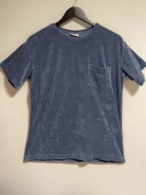 Blå frotté t-shirt från Kappahl - Mysig blå t-shirt från Kappahl i frotté med rund halsringning och en bröstficka. Perfekt för en avslappnad och chill stil. T-shirten har korta ärmar och en enkel, rak passform. Strl: 158-164