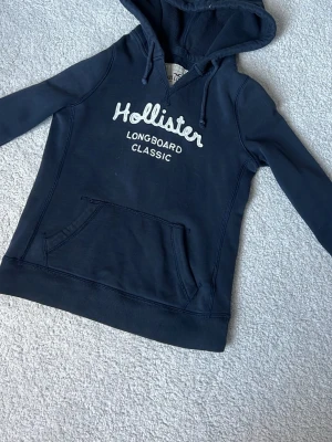 Mörkblå hoodie från Hollister XS - Mörkblå hoodie från Hollister med broderad vit logga och texten 'Longboard Classic' på bröstet. Klassisk känguruficka framtill och huva med dragsko. Perfekt för en avslappnad och cool stil.