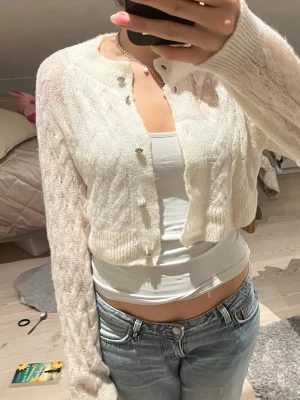 Vit kort kofta med kabelstickning - Supersöt vit kofta i kort modell med kabelstickat mönster och genomskinliga partier. Koftan har rund halsringning, långa ärmar och små pärlknappar framtill. Perfekt att slänga över ett linne för en trendig lager-på-lager-look.