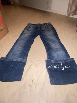 Blåa bootcut jeans med låg midja - Säljer ett par klassiska blåa bootcut jeans med låg midja och slitningar framtill. Jeansen har breda ben och tydliga 2000-talsvibbar, perfekta för dig som gillar retrostil. Flera fickor fram och bak, tillverkade i denim.