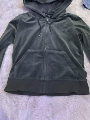 Mörkgrön velour hoodie Juicy Couture - Mörkgrön hoodie i mjuk velour från Juicy Couture med dragkedja och stor ficka framtill. Klassisk huva med snören och snygg J-detalj på dragkedjan. Perfekt för en chill och trendig look.