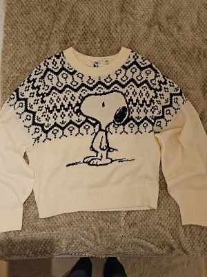 Vit stickad Snoopy-tröja H&M - Stickad tröja från H&M i vitt med svart Snoopy-motiv framtill och grafiskt mönster över axlar och bröst. Rund halsringning och ribbade muddar. Peanuts-kollektion. Perfekt för dig som gillar retro och serietidningsstil.