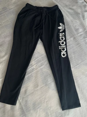 Svarta mjukisbyxor från Adidas S  - Svarta mjukisbyxor från Adidas lös med stort vitt tryck längs ena benet och klassisk logga. Byxorna har snörning i midjan och raka ben. Perfekta för chill eller träning.
