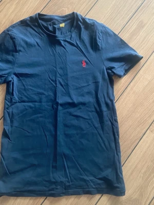 Mörkblå t-shirt från Polo Ralph Lauren - Snygg mörkblå t-shirt från Polo Ralph Lauren med klassisk röd logga broderad på bröstet. T-shirten har rund halsringning och är tillverkad i mjuk bomull. Perfekt för en stilren och avslappnad look.