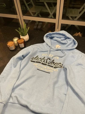 Ljusblå hoodie från Jack & Jones - Ljusblå hoodie från Jack & Jones med stor logga och text på bröstet i vitt och svart. Klassisk huva med dragsko och mjukt bomullsmaterial. Perfekt för en avslappnad och stilren look. Ribbstickade muddar vid ärmslut och nederkant. Våga ställa fler frågor 😁😁