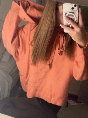 Persikofärgad hoodie med huva - Mysig persikofärgad hoodie med huva och dragsko. Tröjan har en avslappnad passform, ribbade muddar och är tillverkad i ett mjukt stickat material. Perfekt för en chill och trendig look.