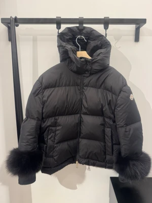 Svart dunjacka med pälskant Moncler - Snygg svart dunjacka från Moncler med stor huva och fluffiga pälskanter vid ärmsluten. Jackan har quiltad design, dragkedja framtill och två fickor. Köptes för 17 000 kr.