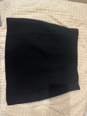 Svart kortkjol från Zara - Snygg svart kortkjol från Zara med stilren design och raka linjer. Kjolen har en klassisk, figurnära passform och är tillverkad i ett mjukt syntetmaterial som känns bekvämt mot huden.