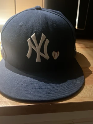 Mörkblå New Era Yankees keps - Snygg mörkblå keps från New Era med New York Yankees-logga framtill och ett litet hjärta som ser ut som en baseboll. Klassisk rak skärm och broderad logga på sidan. Perfekt för dig som gillar sportig streetstyle.