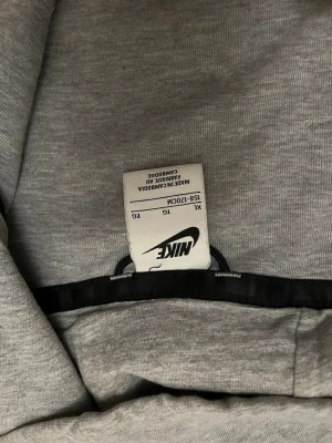 Grå Nike tech hoodie storlek s  - Snygg grå hoodie från Nike i storlek s med hel dragkedja framtill, svart Nike-logga på bröstet och svarta detaljer längs bröstet och ärmslut. Huva och praktisk ficka på ärmen. Perfekt för en sportig och avslappnad stil. Säljer nu för den ha blivit förliten men är i bra skick.
