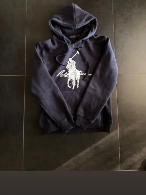 Mörkblå hoodie från Ralph Lauren - Mörkblå hoodie från Ralph Lauren med stor vit logga framtill. Klassisk huva med dragsko och magficka. Mjuk och skön känsla, perfekt för en avslappnad stil. Snyggt tryck med polospelare på bröstet.