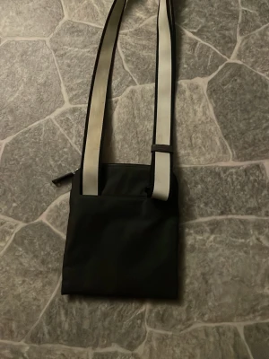 Svart axelväska från Hugo Boss - Snygg svart axelväska från Hugo Boss med vit logga och detaljer framtill. Väskan har en bred, justerbar axelrem i vitt med svarta kanter och dragkedja upptill. Perfekt för att bära det viktigaste och ge en stilren touch till outfiten.