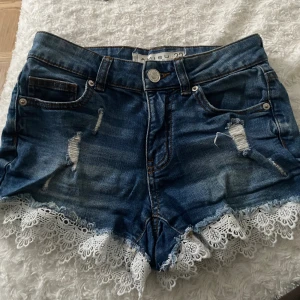 Blå jeansshorts med spets från Amisu - Snygga blå jeansshorts från Amisu med slitna detaljer och vit spetskant längst ner. Storlek 32