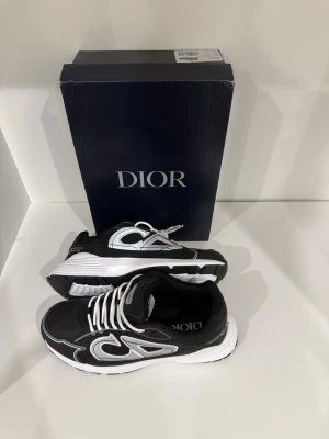 Svarta Dior sneakers med silverdetaljer - Snygga svarta sneakers från Dior med vita och silvriga detaljer. Skorna har en chunky vit och svart sula, tydlig Dior-logga och coola kontraster. Klassisk snörning och sportig design som sticker ut. Perfekta för dig som gillar exklusiva märken och streetstyle.